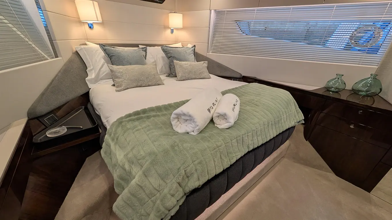 A stylish bedroom with a double bed, green throw, and rolled towels. Features elegant pillows and glass decor on a wooden nightstand.